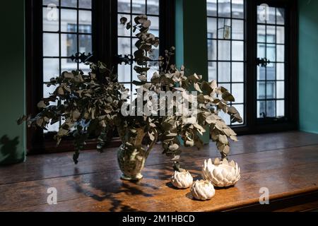 Bouquet of artificial eucalyptus branches in spotted ceramic jug with white porcelain artichoke figurines on wide wooden oak windowsill against the ba Stock Photo