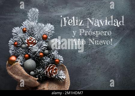 Small Christmas tree in sackcloth decorated with red baubles and berries on dark textured grunge background. Feliz Navidad means Merry Christmas in Stock Photo