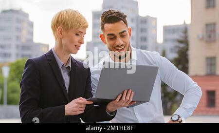 Serious diverse multiracial couple colleagues arabic hispanic man and caucasian woman standing near business center at street outdoors and working on Stock Photo