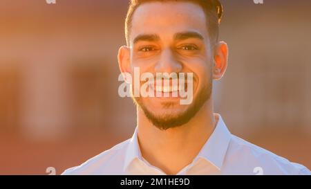 Portrait arabic Hispanic man businessman ethnic bearded guy wears formal shirt stands posing outdoors in street in sun sunset looking at camera Stock Photo
