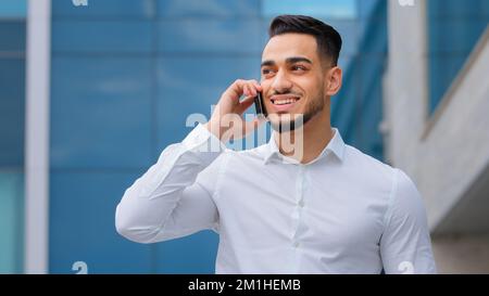 Hispanic indian bearded businessman with mobile phone at street smiling Arabian businessman manager answer call with smartphone outdoors distant Stock Photo