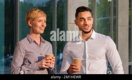 Multiracial colleagues caucasian business woman and hispanic bearded man enjoying break stand outdoors in sunbeams sunset near company building talkin Stock Photo
