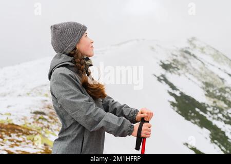 Smiling young woman in freckled hiker in stands on top of a mountain against a backdrop of snow-covered winter mountains, leaning on a hiking pole Stock Photo