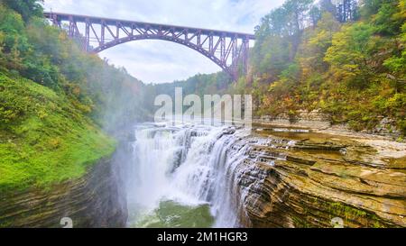 Waterfall cutting through cliffs from drone with steel maroon bridge overhead Stock Photo