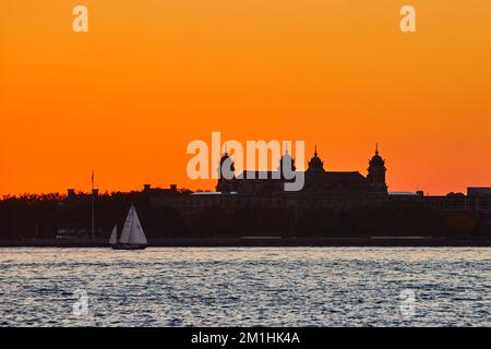 Silhouette of Ellis Island with boat sailing past and intense orange sky Stock Photo