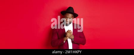 Cunning black man having an idea, smiling devious and rubbing hands, standing against red background and scheming Stock Photo