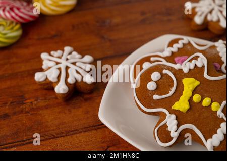Two gingerbread men with colorful icing on a white dessert plate on a wooden table with gingerbread and caramels in the background. Stock Photo