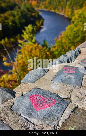 Detail stone wall with pink heart chalk art and soft river with fall forest behind Stock Photo