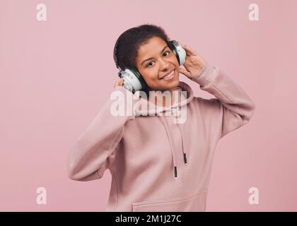 Happy beautiful young woman listening music in headphones. Charming black girl in hoodie and big white earphones dancing against pink background Stock Photo