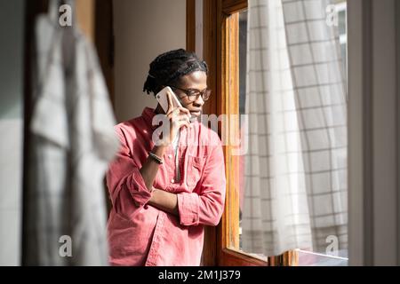 Sad upset African man having difficult conversation on phone, standing by window at home Stock Photo