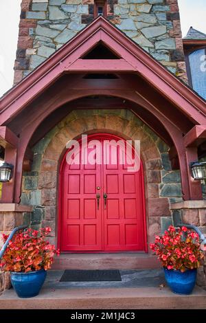 Straight on pair of red arched doors with stone wall and red plants Stock Photo