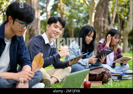 Handsome and smart young Asian male college student using his portable tablet while sitting in the university's park with his friends. outdoor learnin Stock Photo