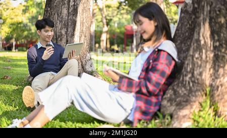 Handsome and cheerful young Asian male college student using his portable tablet and sipping coffee while relaxing in the park with his friend. Stock Photo