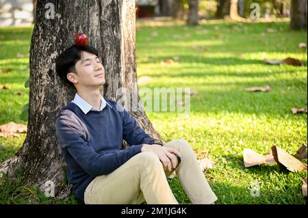 Relaxed young Asian man in casual clothes sitting under the tree, eyes closed, chilling at the beautiful greenery park. Stock Photo