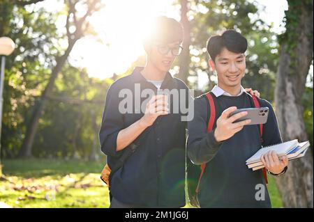 Two young Asian men college students looking at smartphone screen while walking in the campus's park together. Stock Photo