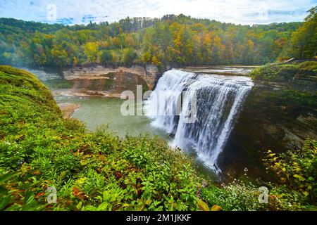 Huge waterfall wide angle up close pouring over cliffs into canyon in lush green forest with hints of approaching fall Stock Photo