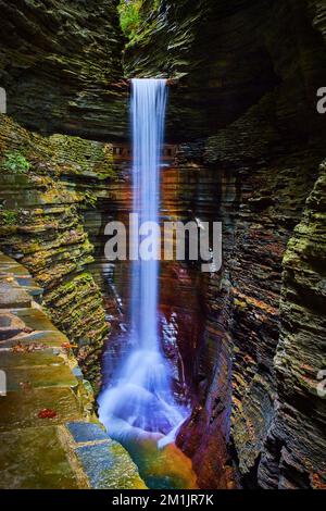 Tall waterfall pours into deep gorge of layered rocks and path leading behind Stock Photo