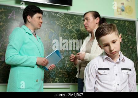 Sad upset schoolboy against the background of talking mom and teacher. Problems at school. The teacher complains to the mother about the student. Stud Stock Photo