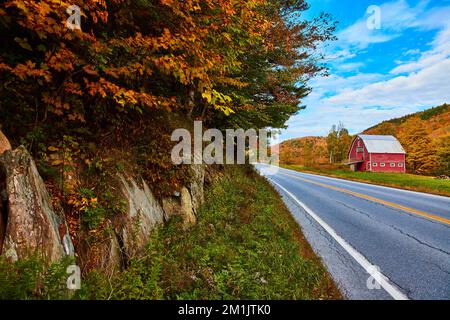 Roadside view of rocky walls, peak fall foliage hills, and stunning red barn in background Stock Photo