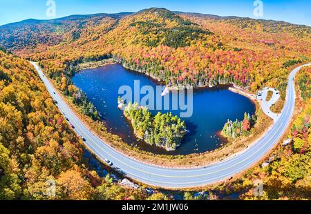 Wide panorama aerial over highway through stunning peak fall foliage New Hampshire mountains around blue lake Stock Photo