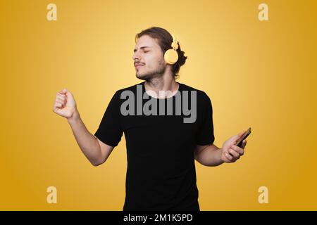 Young man wearing headphones having fun listening favorite music while keeping eyes closed isolated against yellow background. Mobile app. Happy Stock Photo