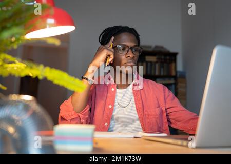 Tired confused African American man looking at laptop screen sits at office table with notepad Stock Photo