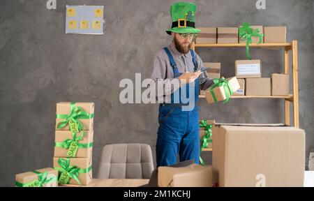 male warehouse worker seller in leprechaun hat, small stock business owner holding smartphone, using mobile app scanning retail package parcel bar cod Stock Photo