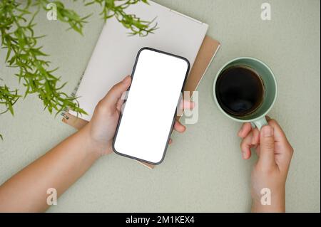 Top view, A female hands holding a smartphone white screen mockup and a cup of coffee on her modern desk. Stock Photo