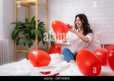 Girl is blowing air balloon at home Handsome brunette girl inflating red heart shaped balloon sitting on bed. Preparation for valentines day party Stock Photo