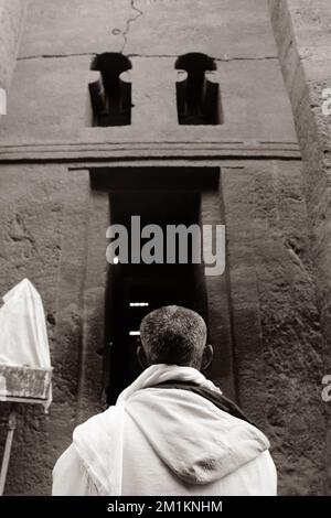 A grayscale vertical shot of a man standing in front of a church entrance in Lalibela, Ethiopia Stock Photo