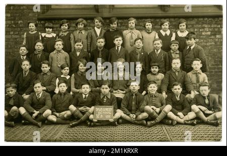 Original 1920's era postcard of Brae Street School V1B dated 1928 (on sign)  group class portrait outside in the school playground, wearing mixed school uniforms and other fashions. On reverse is written age 12 years. Liverpool, Merseyside, England, U.K. Stock Photo