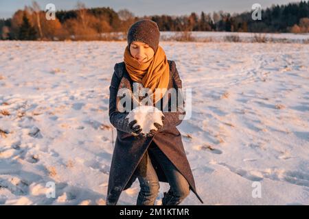 A woman in winter clothes collects a lump of snow in a snow-covered field Stock Photo