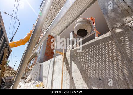 Shot from bellow on yellow long plastic tube placed next to building under construction for throwing industrial waste in smeared tipper truck trailer. Stock Photo