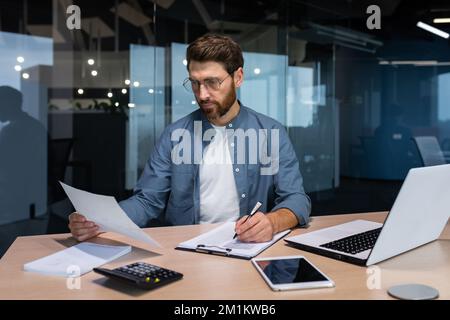 Serious and focused financier accountant on paper work inside office, mature man using calculator and laptop for calculating reports and summarizing accounts, businessman at work in casual clothes. Stock Photo