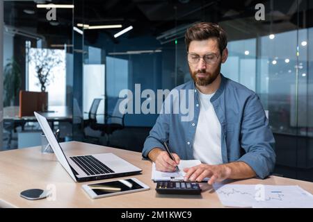 Serious and focused financier accountant on paper work inside office, mature man using calculator and laptop for calculating reports and summarizing accounts, businessman at work in casual clothes. Stock Photo
