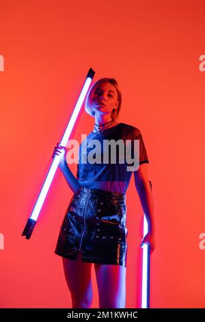 low angle view of woman in leather mini skirt holding fluorescent lamps on coral background,stock image Stock Photo