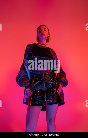 low angle view of woman in leather mini skirt looking at camera in neon light on coral and pink background,stock image Stock Photo
