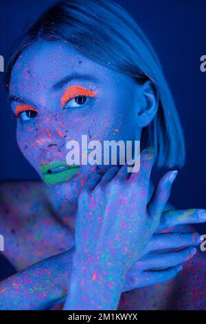 woman with glowing makeup and paint splashes posing in neon light isolated on dark blue,stock image Stock Photo