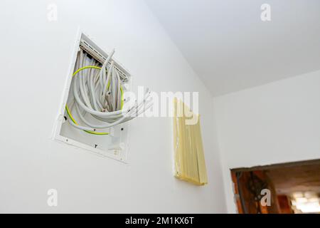 Bundle of various cables hang from an open plastic fuse switch box mounted on the plaster wall, electric power meter covered with duct tape installed Stock Photo
