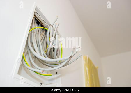 Bundle of various cables hang from an open plastic fuse switch box mounted on the plaster wall, electric power meter covered with duct tape installed Stock Photo