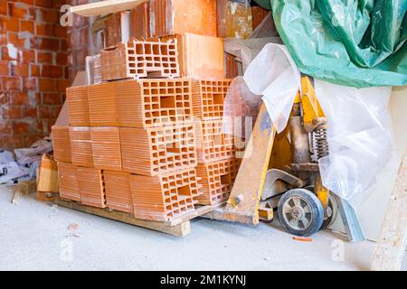Stacked red blocks at the wooden pallet placed on metal, manually forklift in warehouse of construction site. Stock Photo