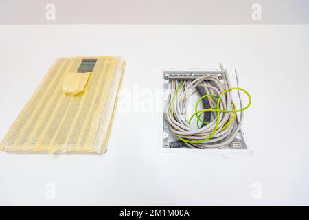 Bundle of various cables hang from an open plastic fuse switch box mounted on the plaster wall, electric power meter covered with duct tape installed Stock Photo