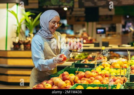 Female seller in hijab browsing and checking apples in supermarket, woman in apron smiling at work in store in fruit and vegetable department. Stock Photo