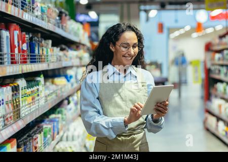 Latin American woman seller happy and smiling uses tablet computer in supermarket to check stock availability, shop assistant among shelves with goods in apron. Stock Photo