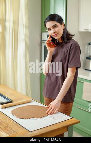 Vertical portrait of young mixed race woman talking on smartphone in home kitchen while cooking, look at camera Stock Photo
