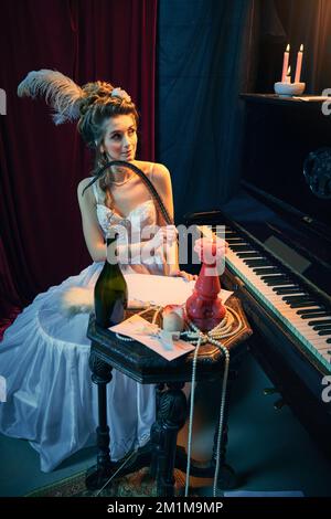 Portrait of young beautiful girl in image of medieval person in elegant white dress sitting at the piano and writing novel. Comparison of eras, beauty Stock Photo