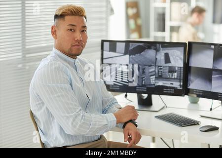 Side view portrait of young Asian man looking at camera with surveillance feeds in background, security center office, copy space Stock Photo