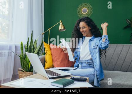 Happy hispanic woman at home doing paperwork and calculating household budget, sitting on couch in living room and using laptop at work, holding hand up victory and triumph gesture. Stock Photo