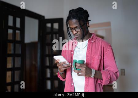 Young African man holding cup of tea and smartphone texting message while spending free time at home Stock Photo