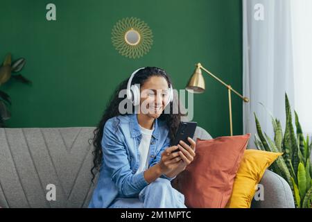 Young smiling hispanic woman at home with headphones and phone listening to music online using smartphone app sitting on sofa in living room and dancing. Stock Photo